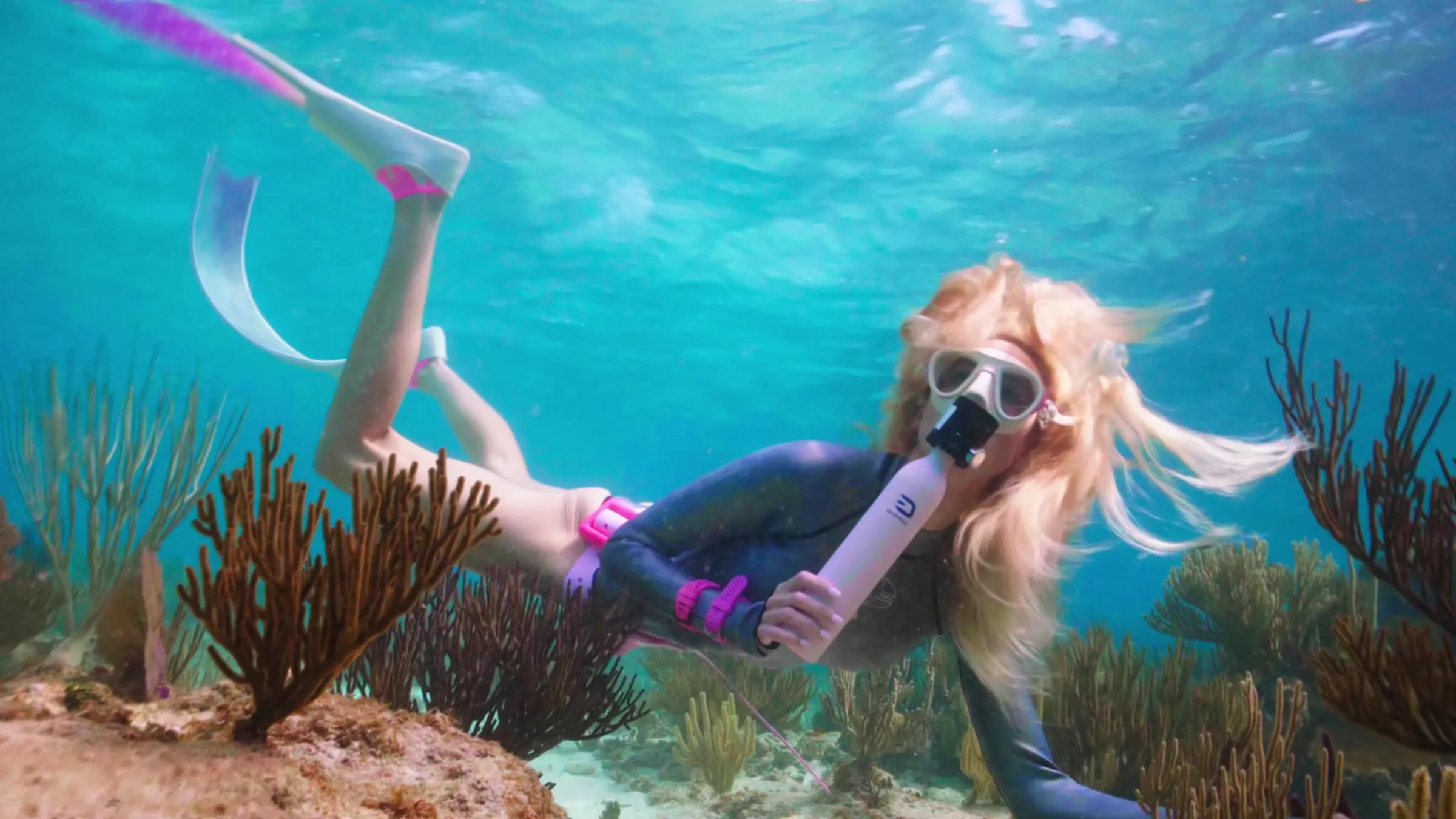 A blonde woman with a mask and pink fins swims over orange sea fans, breathing from a small pink Goyojo tank held to her mouth.