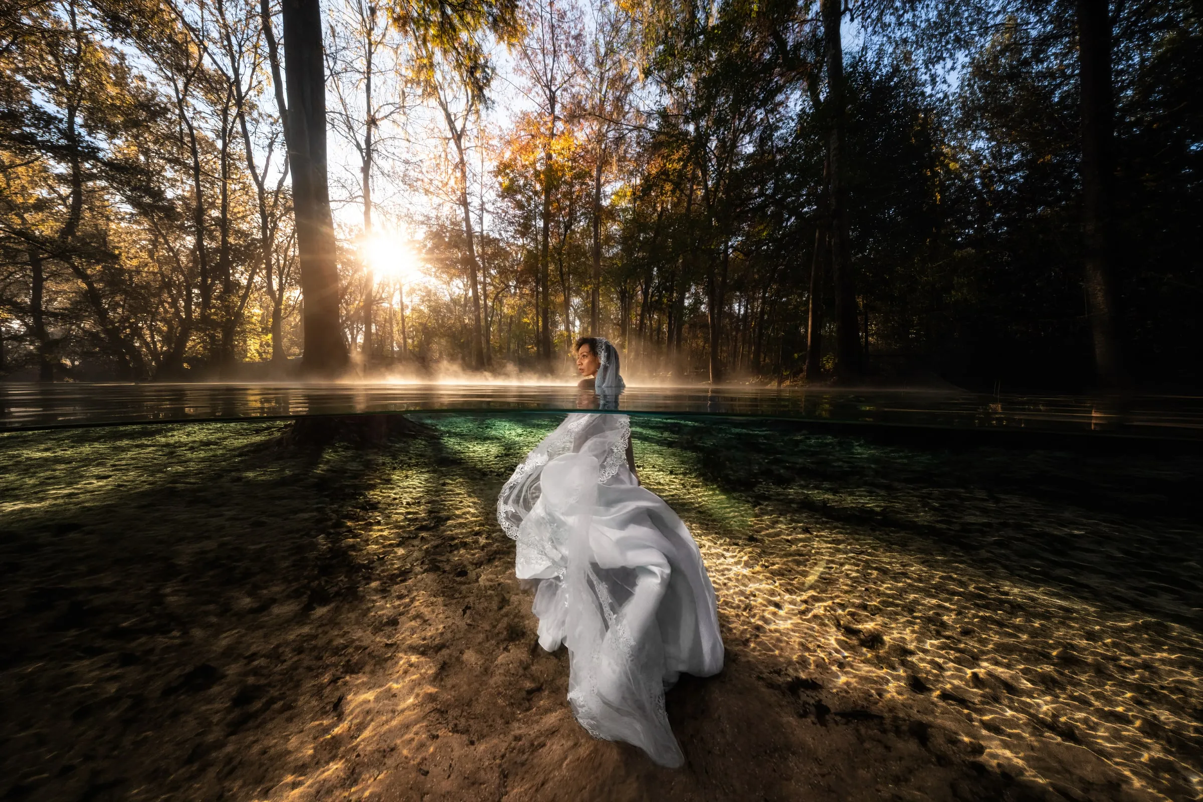 A bride in a lace-edged white gown and veil stands in a clear spring with only her head above the water, low sun and autumn trees behind her.