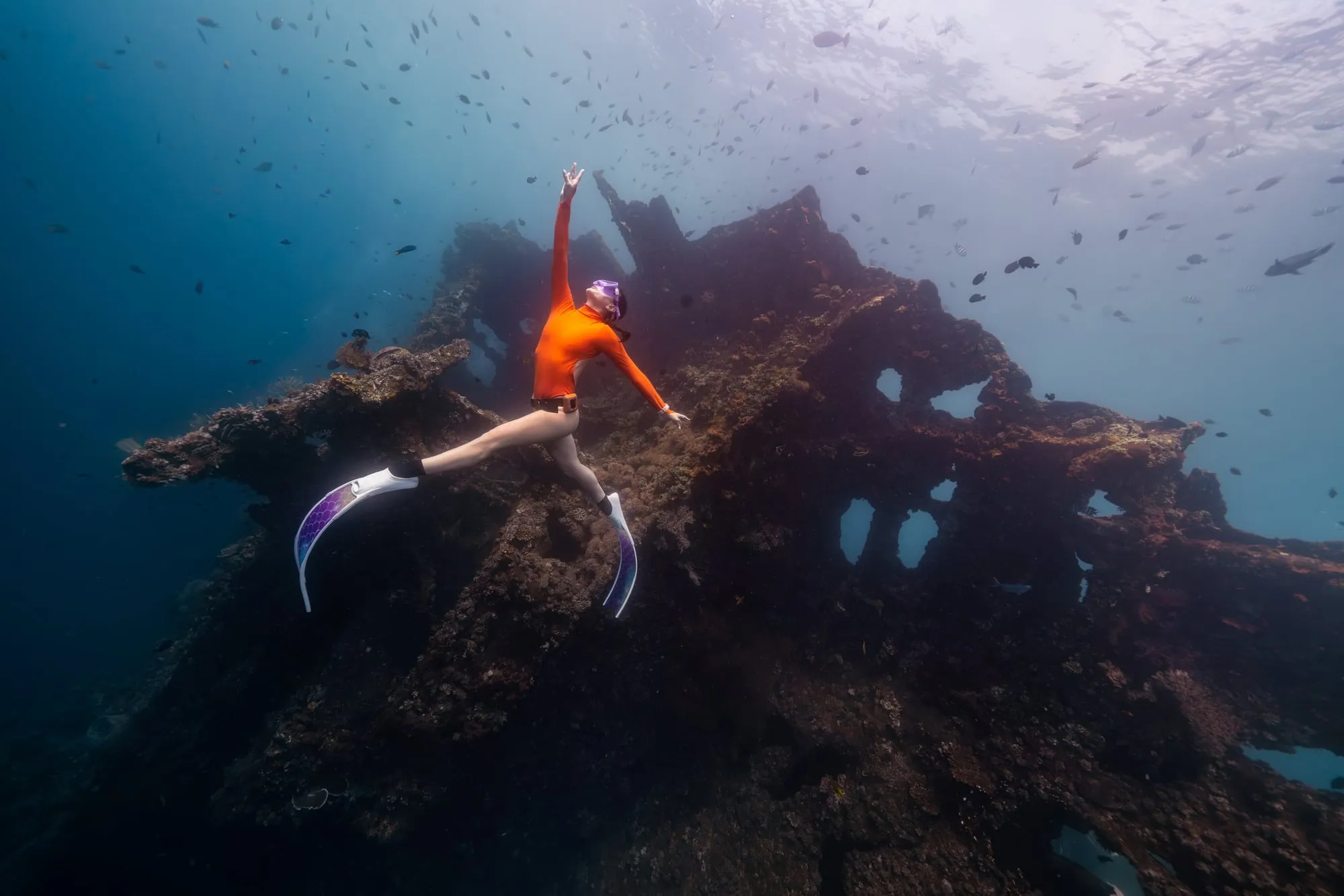 A freediver in an orange top and long white fins reaches up from the bow of a sunken wreck, fish drifting in the blue water around her.