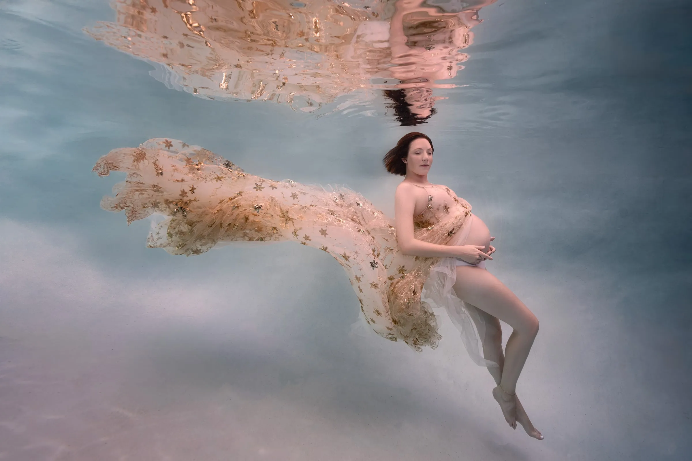 An expectant mother in a gold-flecked tulle gown lies on her side in pale water, the fabric trailing out to her left and her reflection carried along the surface above.