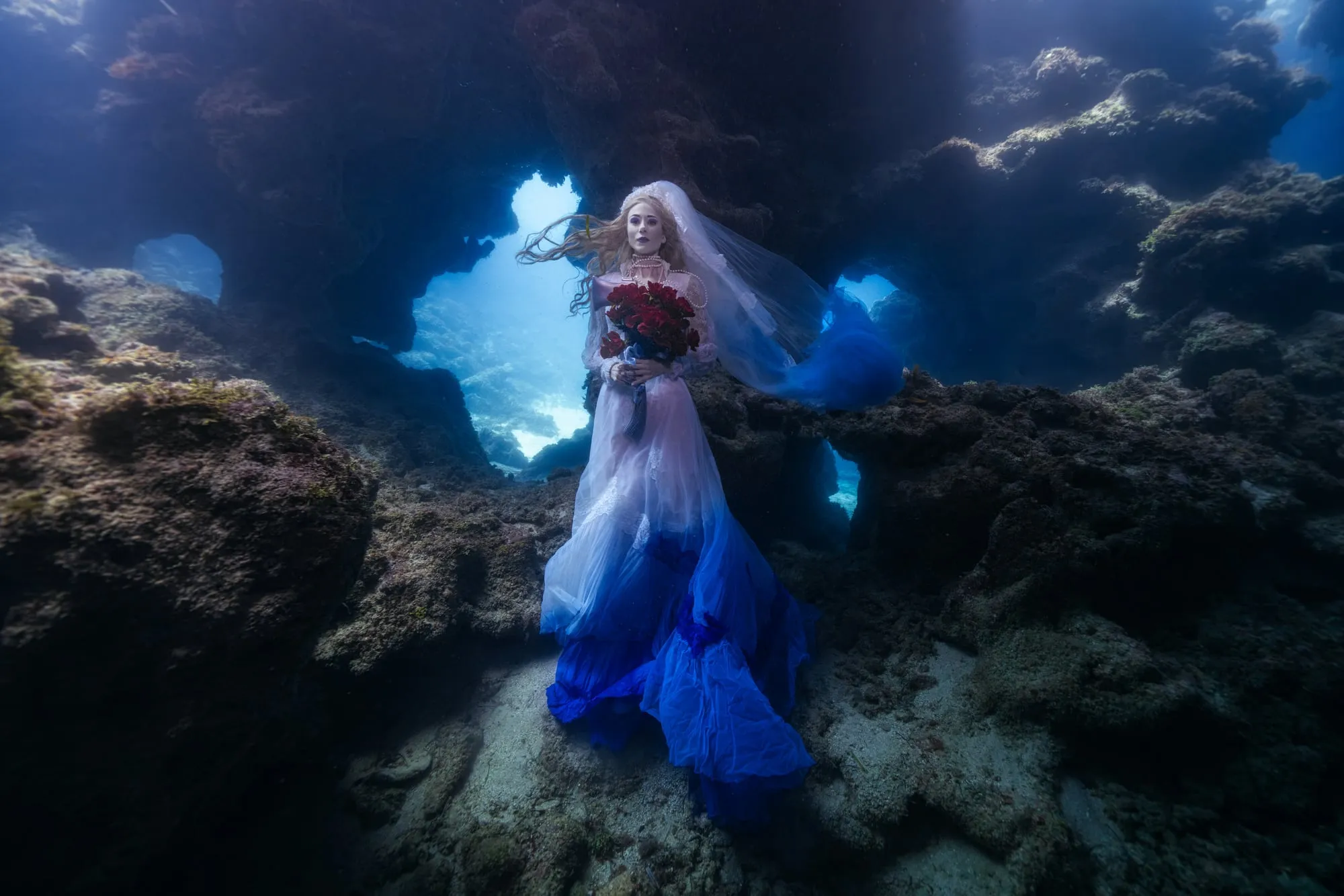 A woman in a white veil and a blue train stands in the lit opening of a cave, holding red roses, rock walls around her.