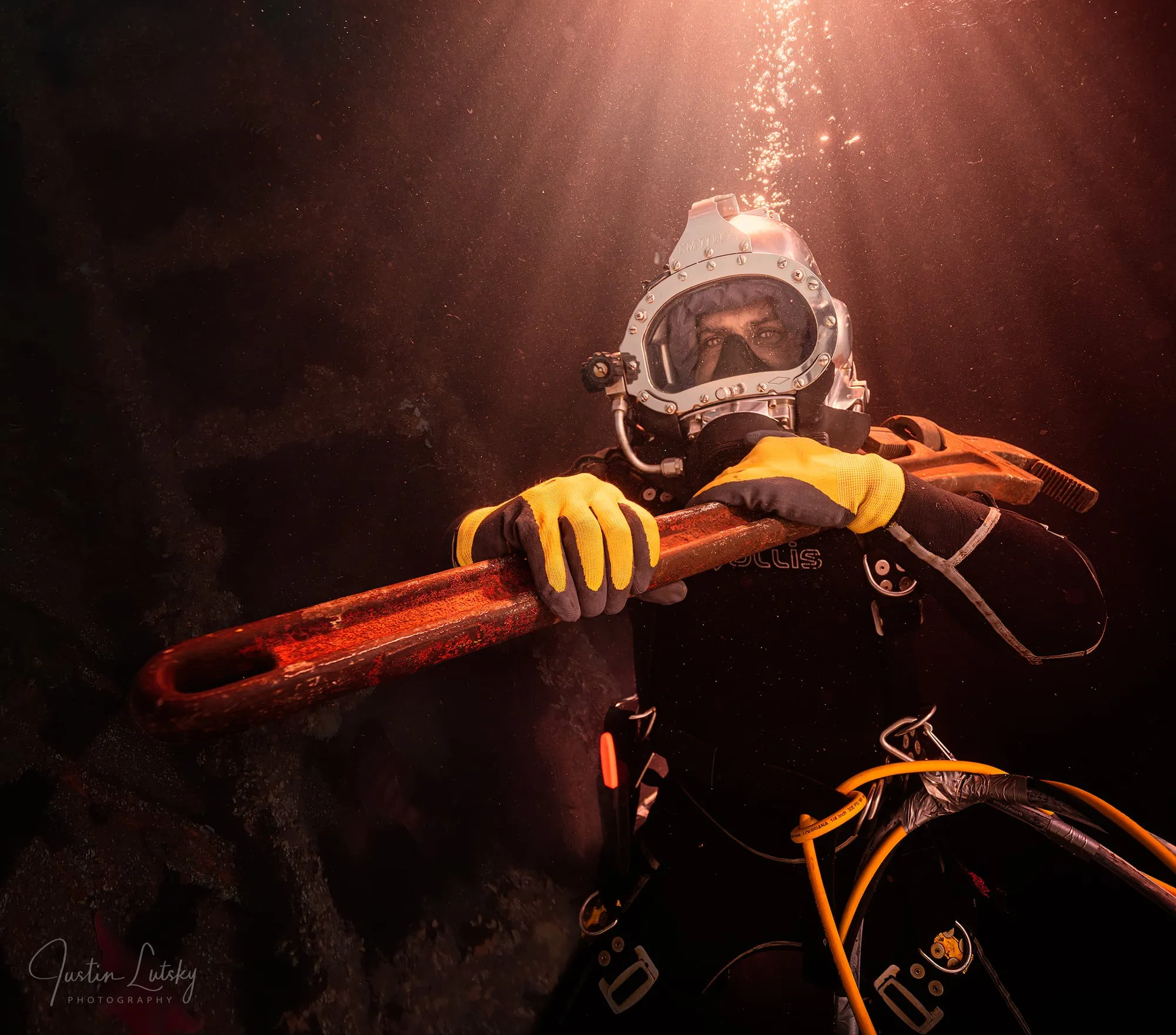 A diver in a vintage helmet and wetsuit grips an orange wrench, lit warm from above, bubbles rising through dark water.