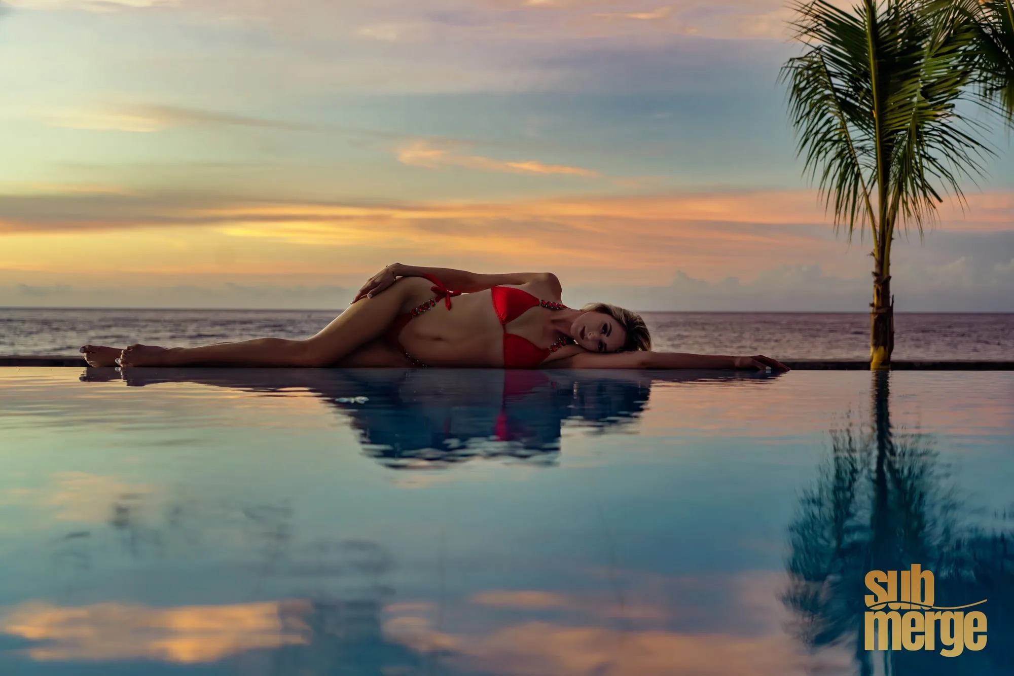 A woman in a white swimsuit leans back in the shallows of a palm-fringed beach, one arm over her head.