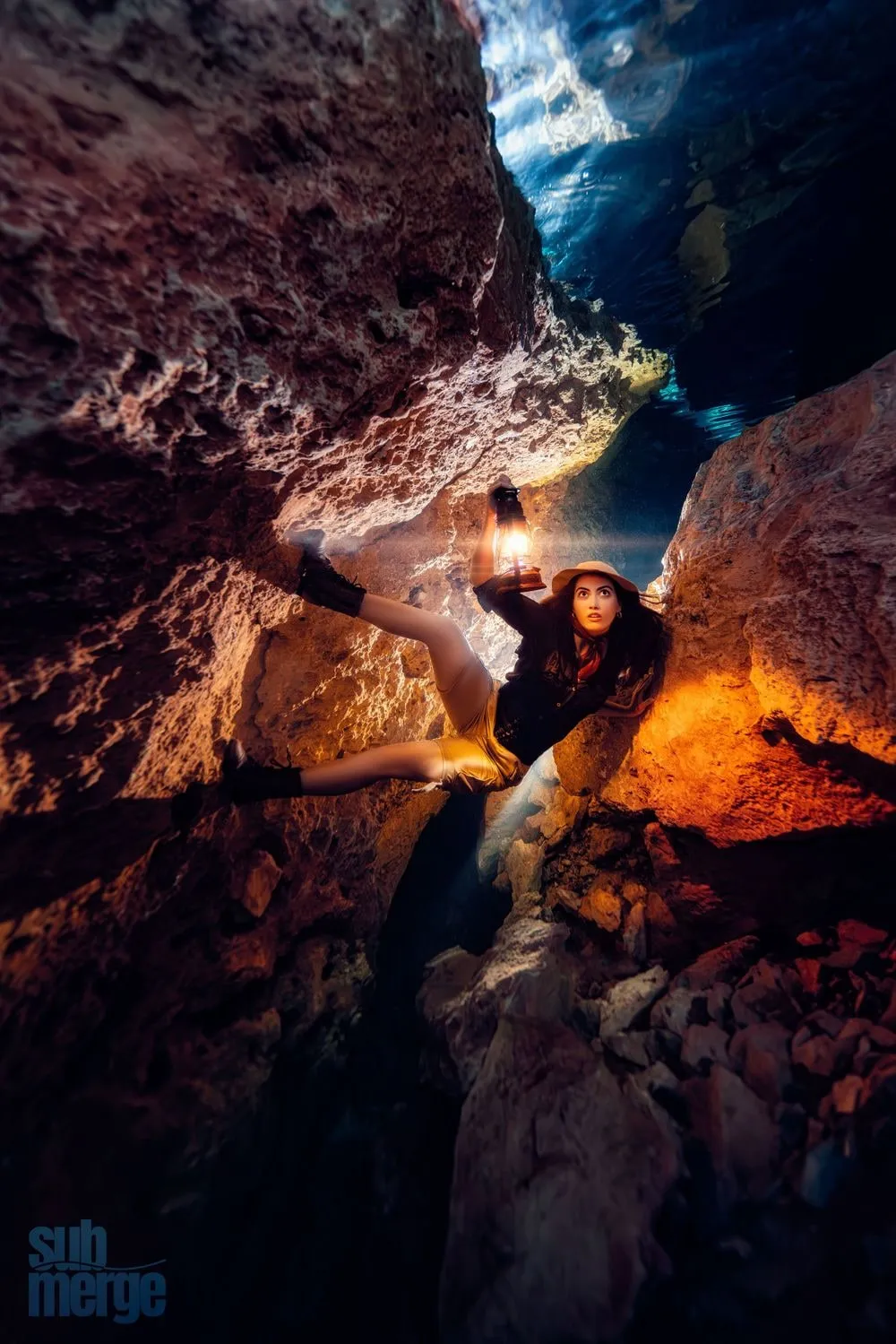 A model in explorer costume braces between rock walls in a flooded cave, holding a lit oil lantern.