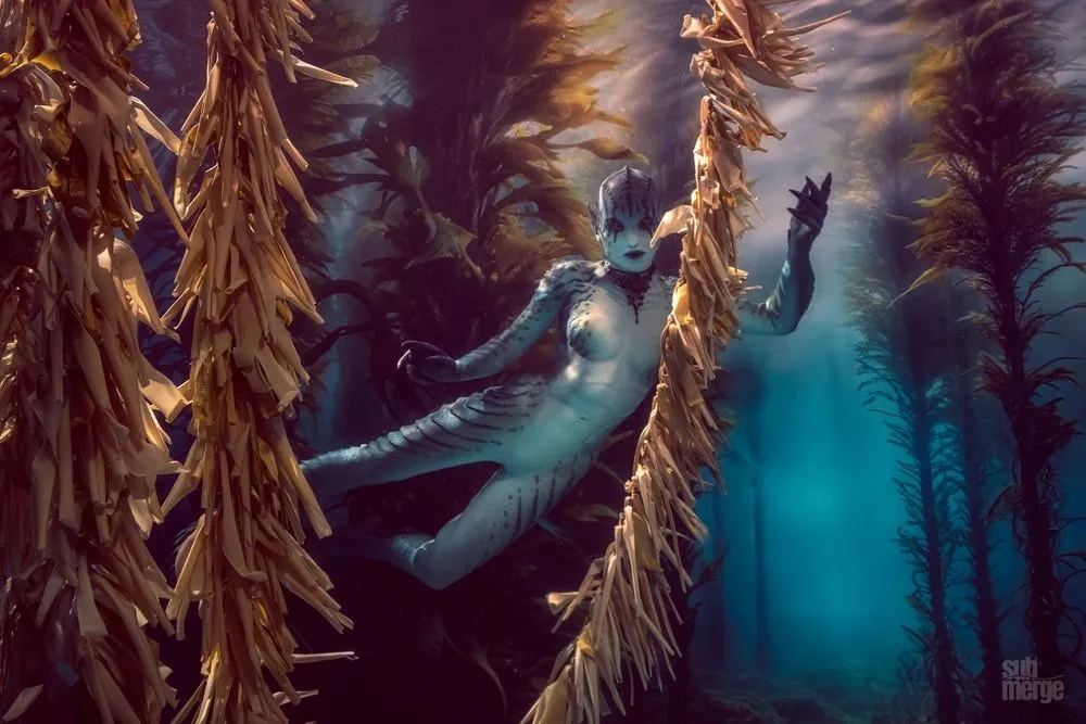 A performer in full blue-gray creature prosthetics and fins glides through a golden kelp forest.