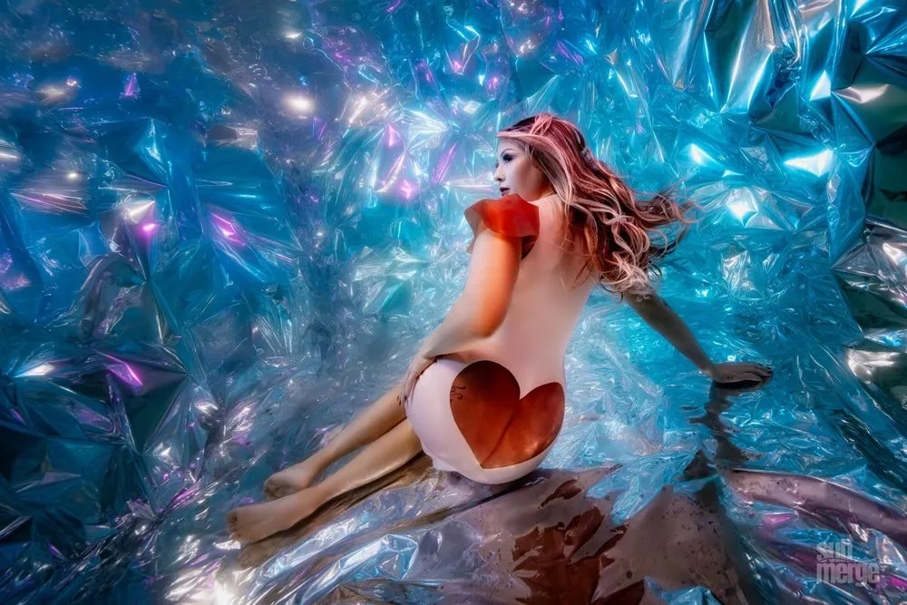 A model poses inside a studio set lined with crumpled iridescent film lit in blue and magenta, a red heart shape across her back.
