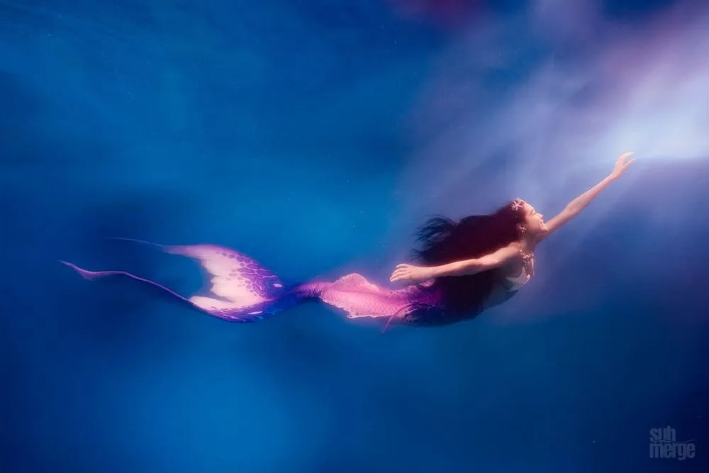 A mermaid in a pink and purple tail arcs through deep blue water, arms outstretched toward a shaft of surface light.