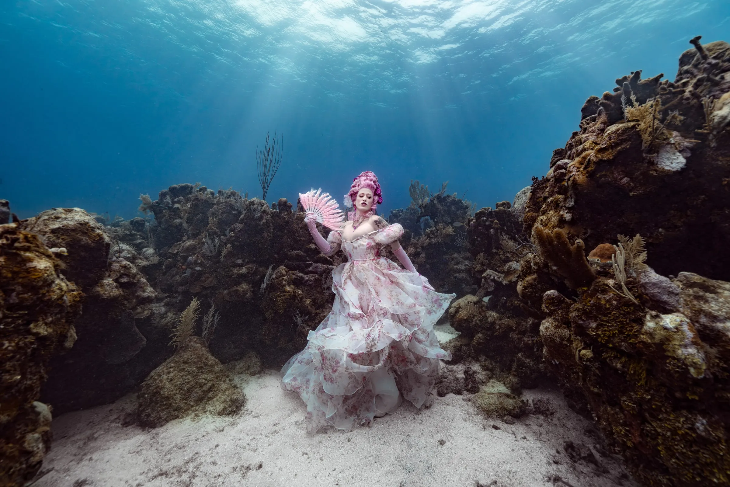A woman in a pink floral organza ballgown and a tall pink wig holds a feathered fan on a sandy floor between two reef walls, sunbeams falling through the water above her.