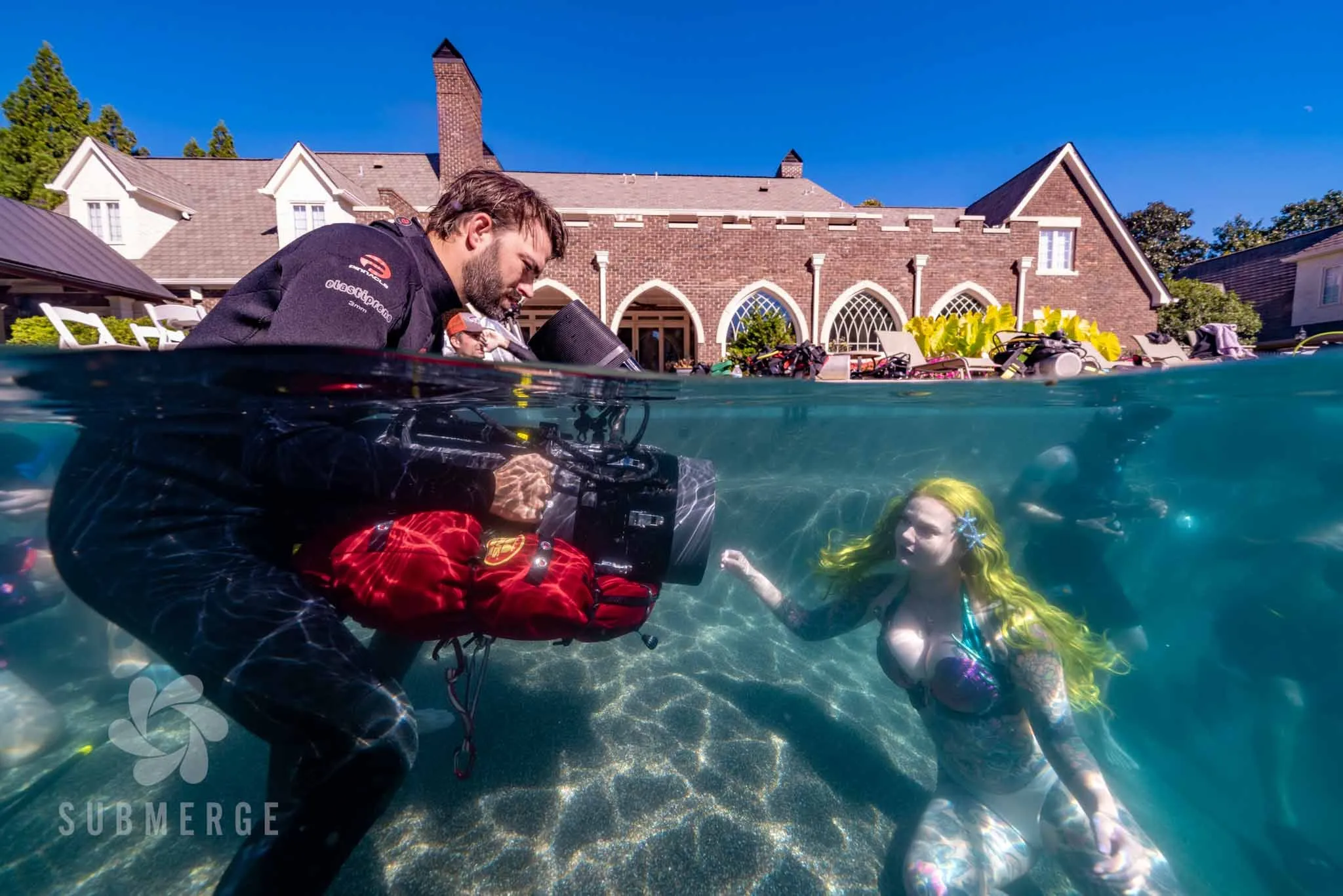 A camera operator at the edge of a pool films a yellow-haired performer beneath the surface, crew and a mansion behind them.