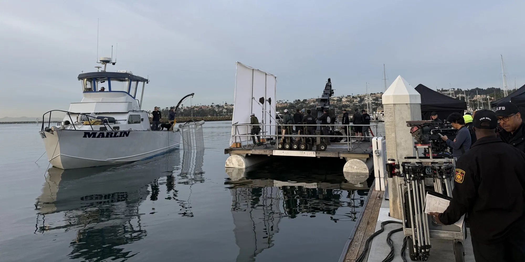 The Marlin, a white 43-foot dive support vessel, tied up at a Los Angeles dock at dawn beside a floating camera platform, a film crew and a crane on its deck and camera carts on the pier.