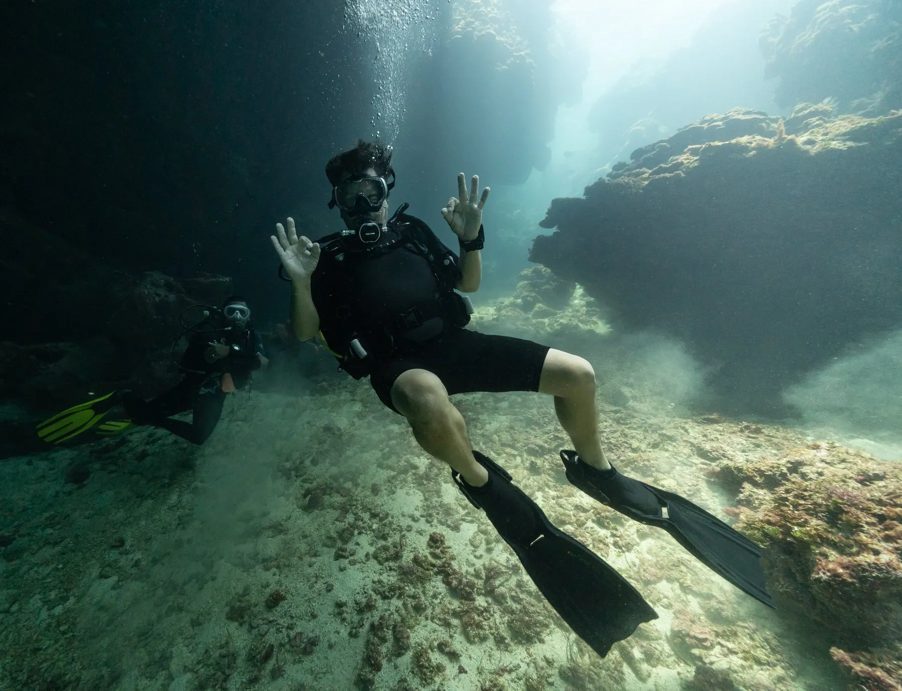An instructor and a student diver kneel on white sand in clear blue water, the instructor signalling OK.