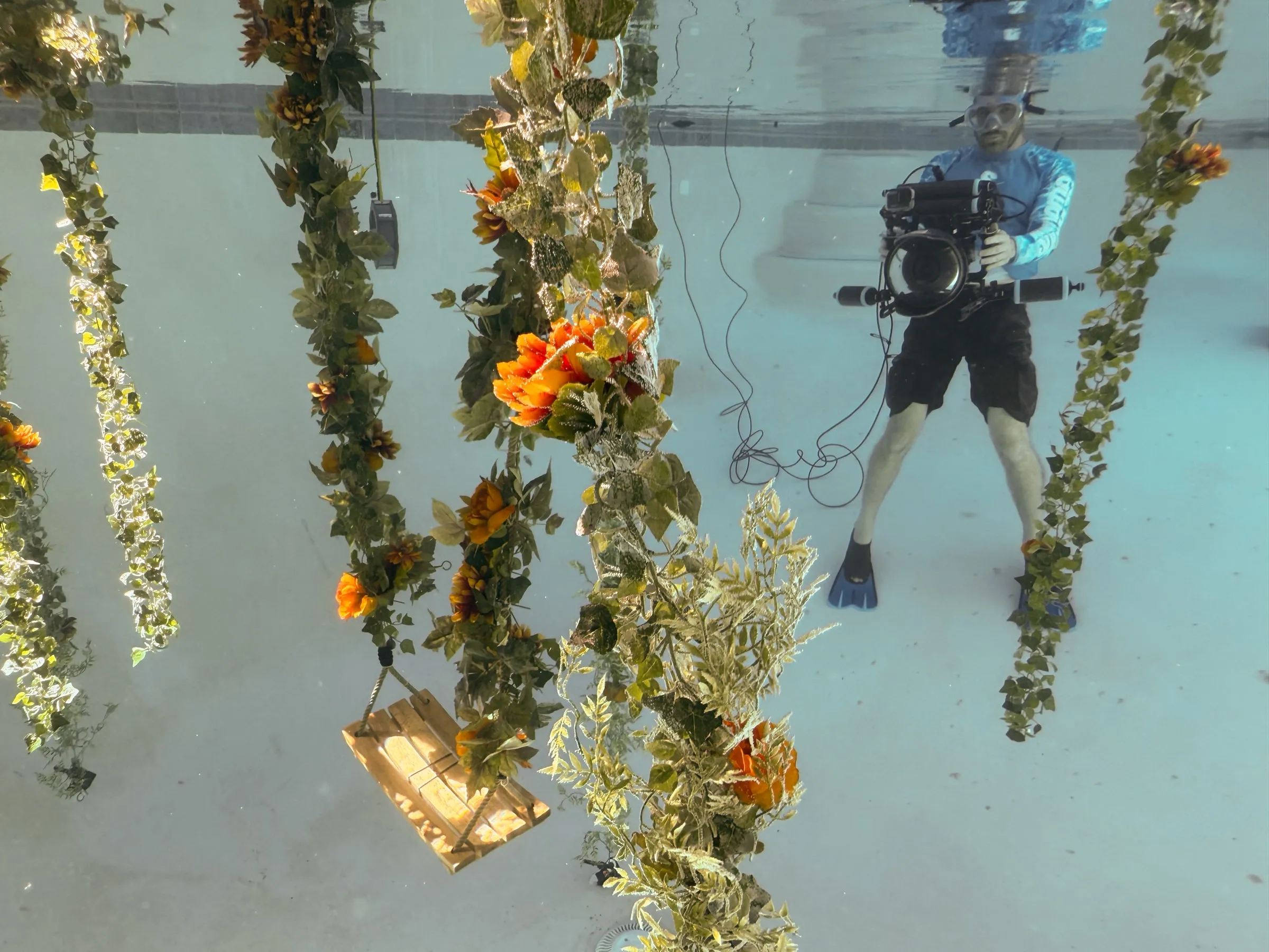 A camera operator in a blue rash guard stands among hanging flower garlands and a wooden swing in the studio pool, a housed camera in his hands.