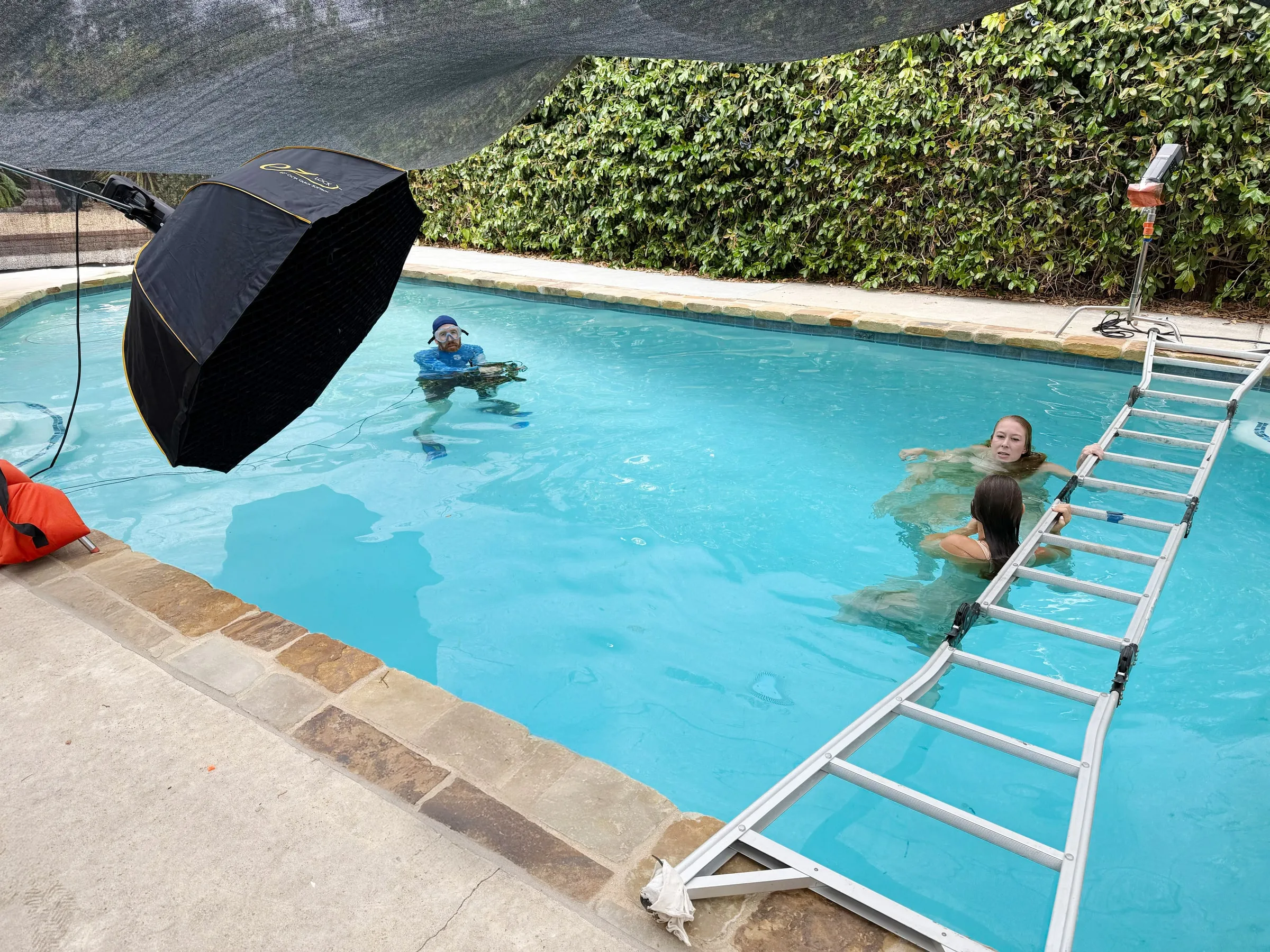 Between frames in the studio pool: the photographer waits in the water with the camera while two women rest on a ladder laid across the pool.