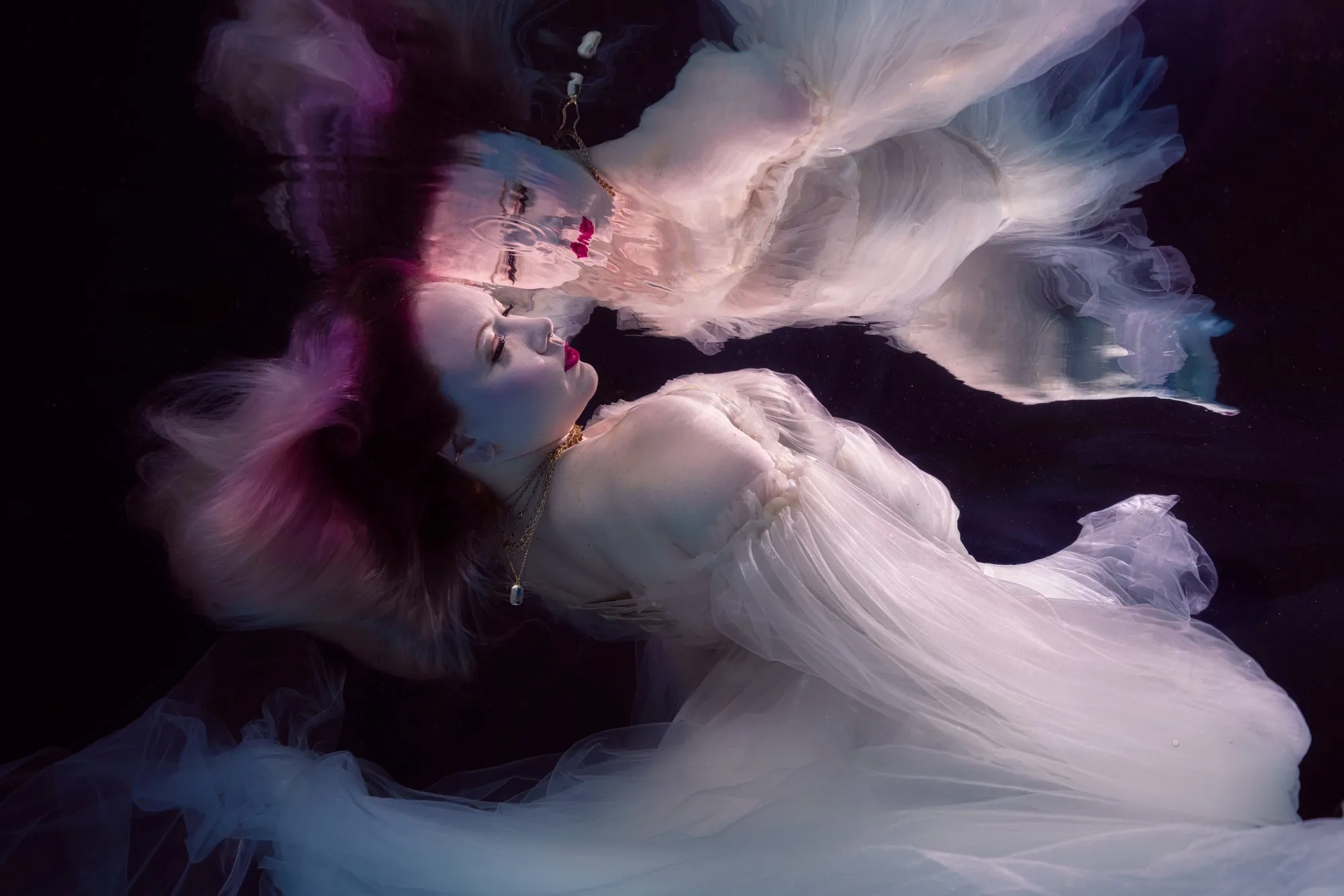 A woman with pink-tinted hair in a white tulle gown floats on her back in dark water, the fabric spreading out around her and her reflection on the surface above.