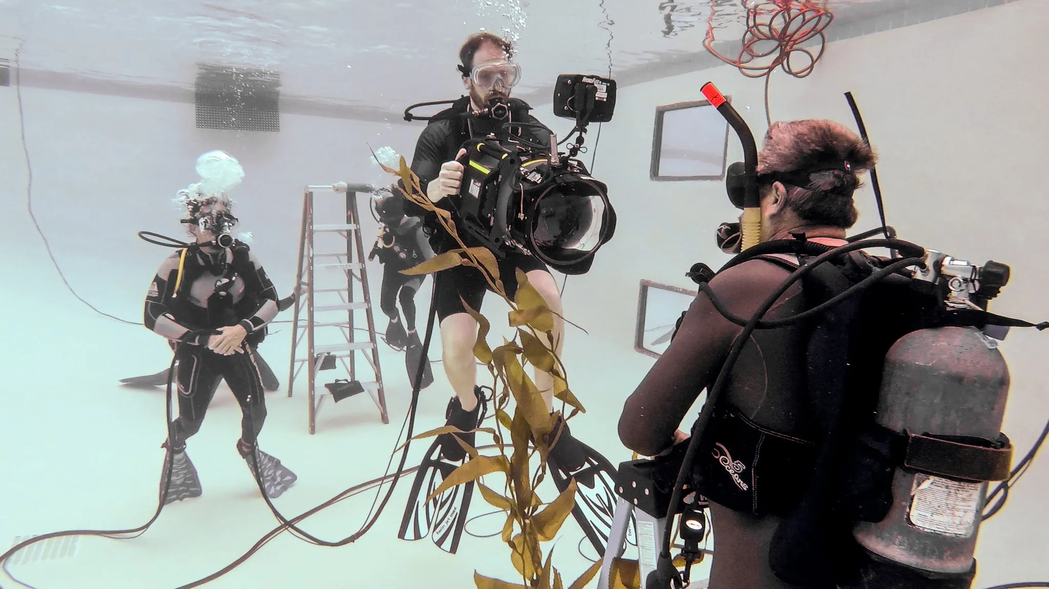 A camera operator holds a housed cinema camera in a white-walled pool, a safety diver and a ladder behind him and a strand of kelp in the foreground.