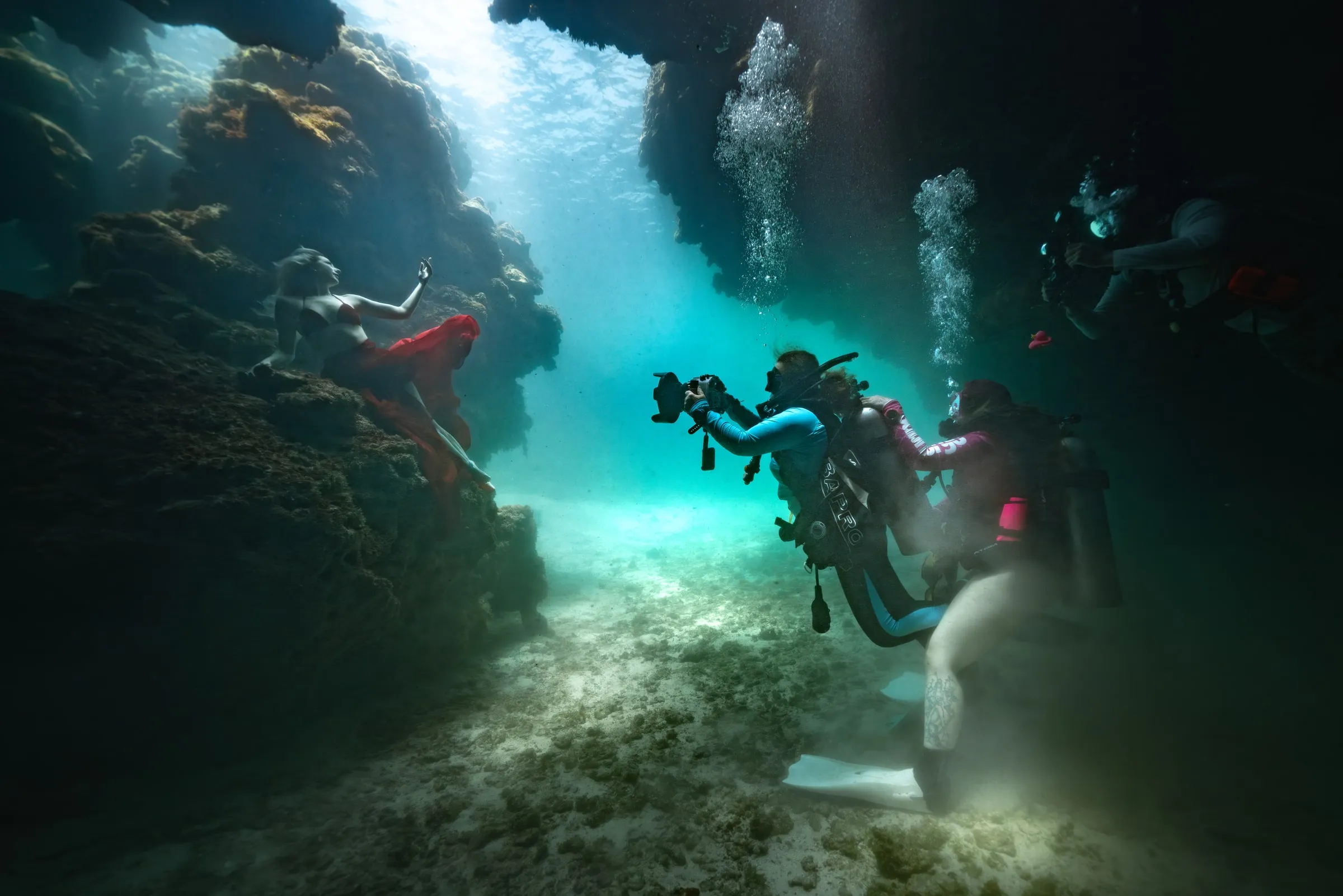 Photographers and safety divers in scuba gear work around a model in the green light of a reef cavern.
